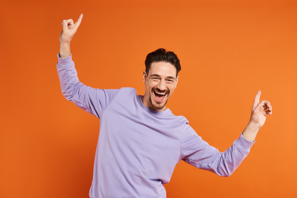 fröhlicher und bärtiger Mann in lila Sweatshirt, der mit den Händen nach oben auf leuchtend orangefarbenem Hintergrund tanzt - Foto, Bild