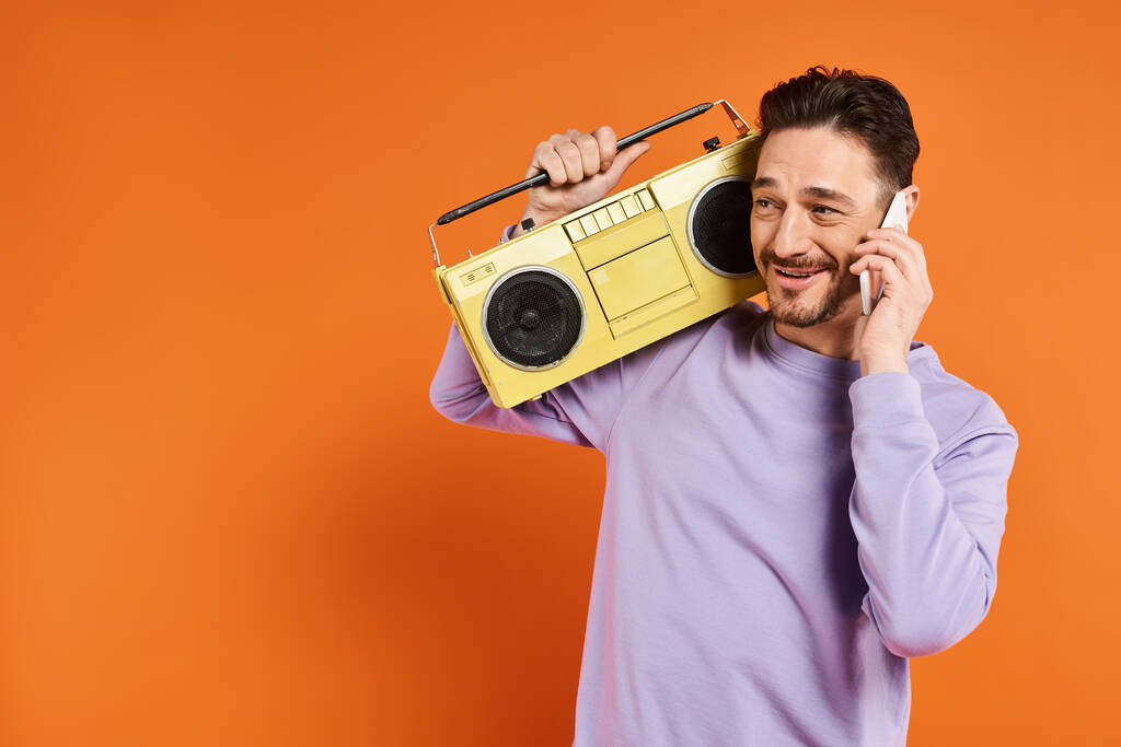 радісний і бородатий чоловік розмовляє на смартфоні і тримає ретро бумбокс на помаранчевому фоні - Фото, зображення
