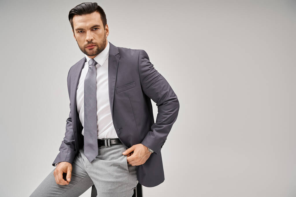 homme d'affaires beau et confiant en tenue élégante assis sur un tabouret haut sur fond gris - Photo, image
