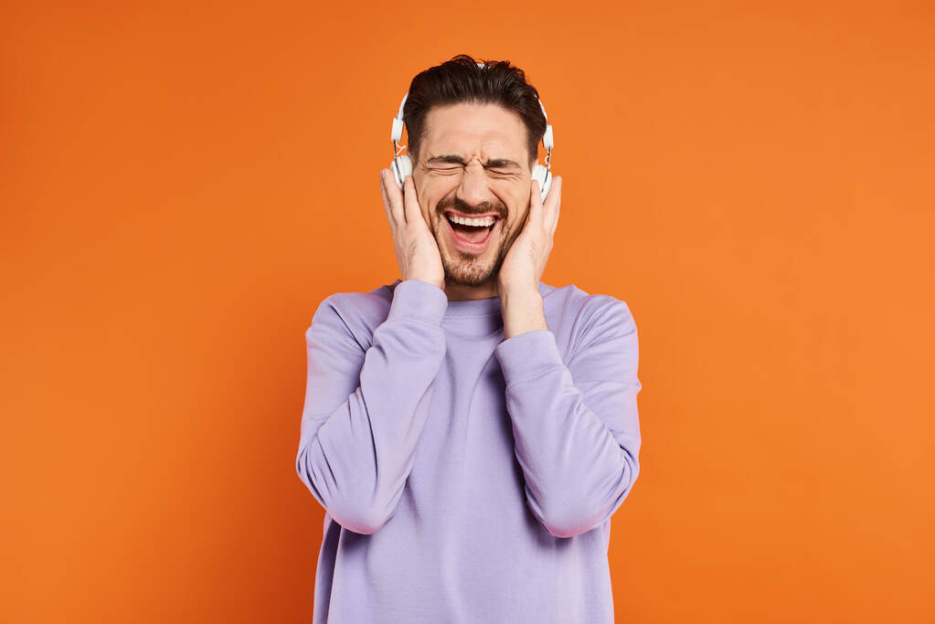 opgewonden bebaarde man in paarse trui en draadloze hoofdtelefoon luisteren muziek op oranje achtergrond - Foto, afbeelding