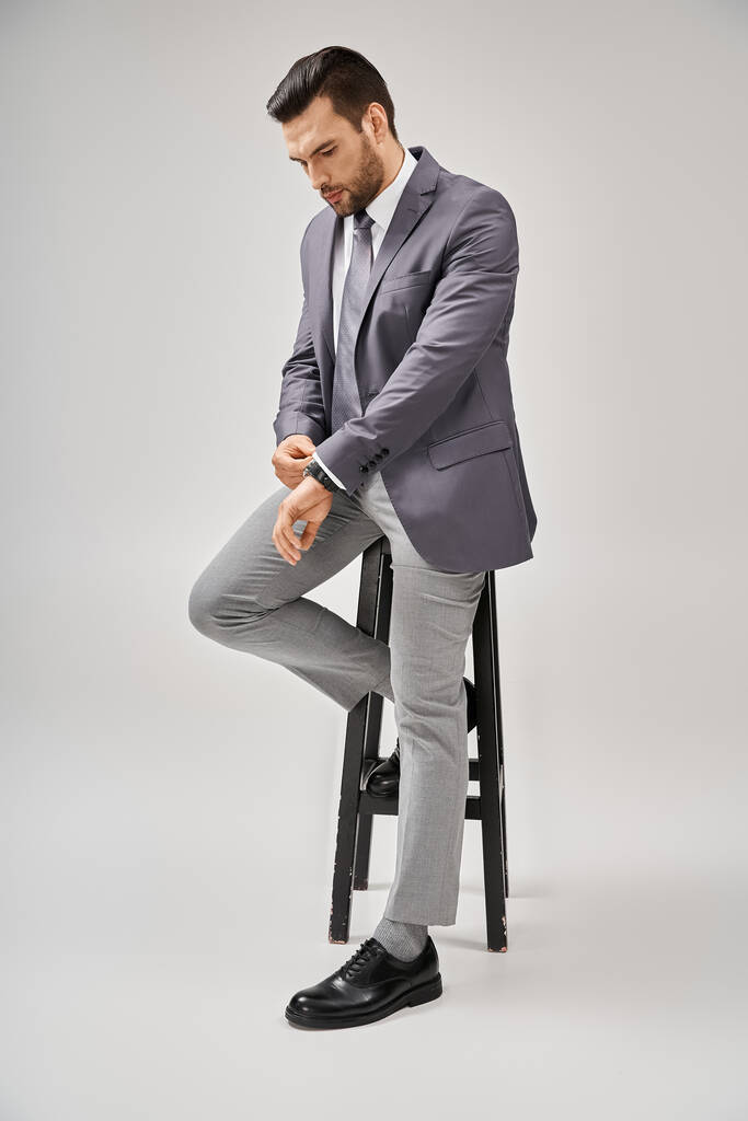 pohledný a sebevědomý podnikatel v obleku opírající se o vysokou stoličku na šedém pozadí, vedení - Fotografie, Obrázek