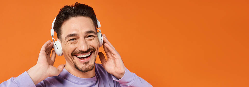 uomo positivo in maglione viola e cuffie wireless ascoltare musica su sfondo arancione, banner - Foto, immagini