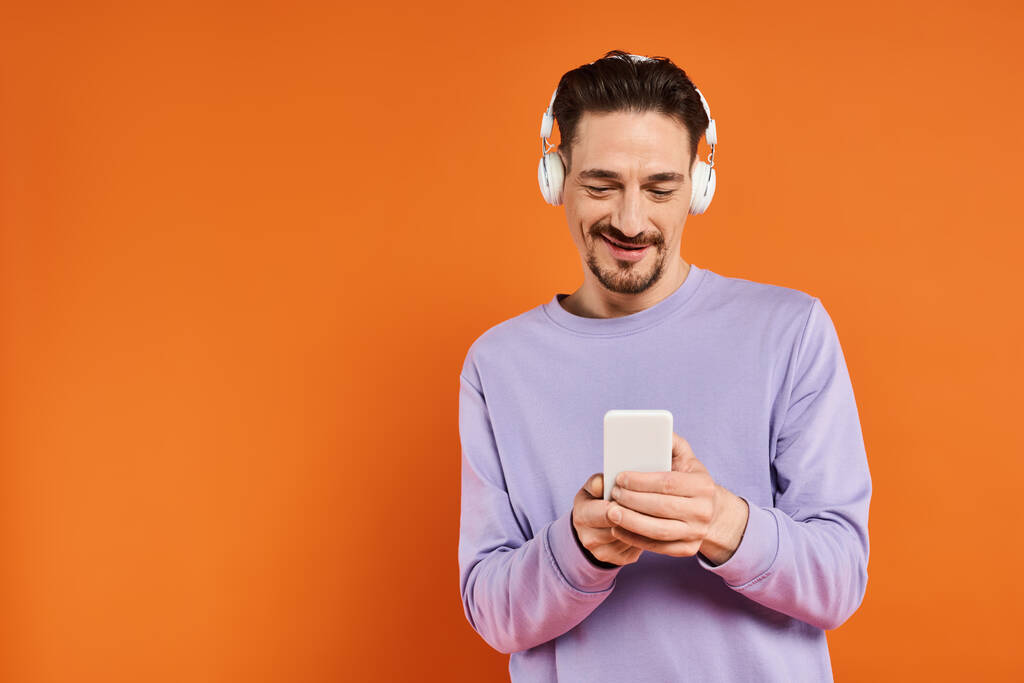 positive man in wireless headphones listening music and using smartphone on orange background - Photo, Image