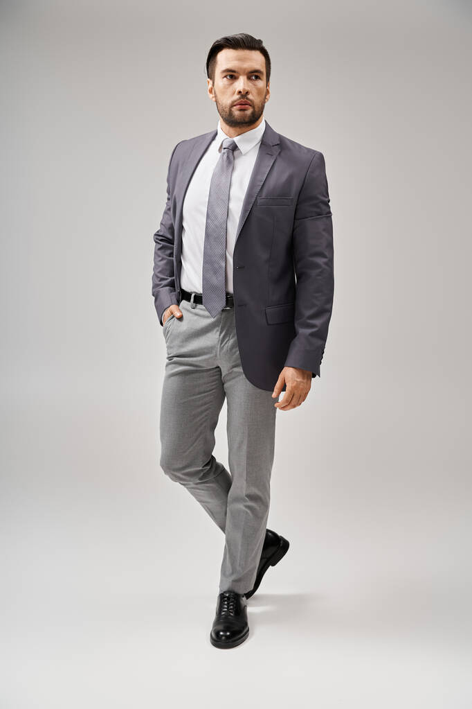 handsome businessman with bristle posing with hand in pocket of pants on grey background - Photo, Image