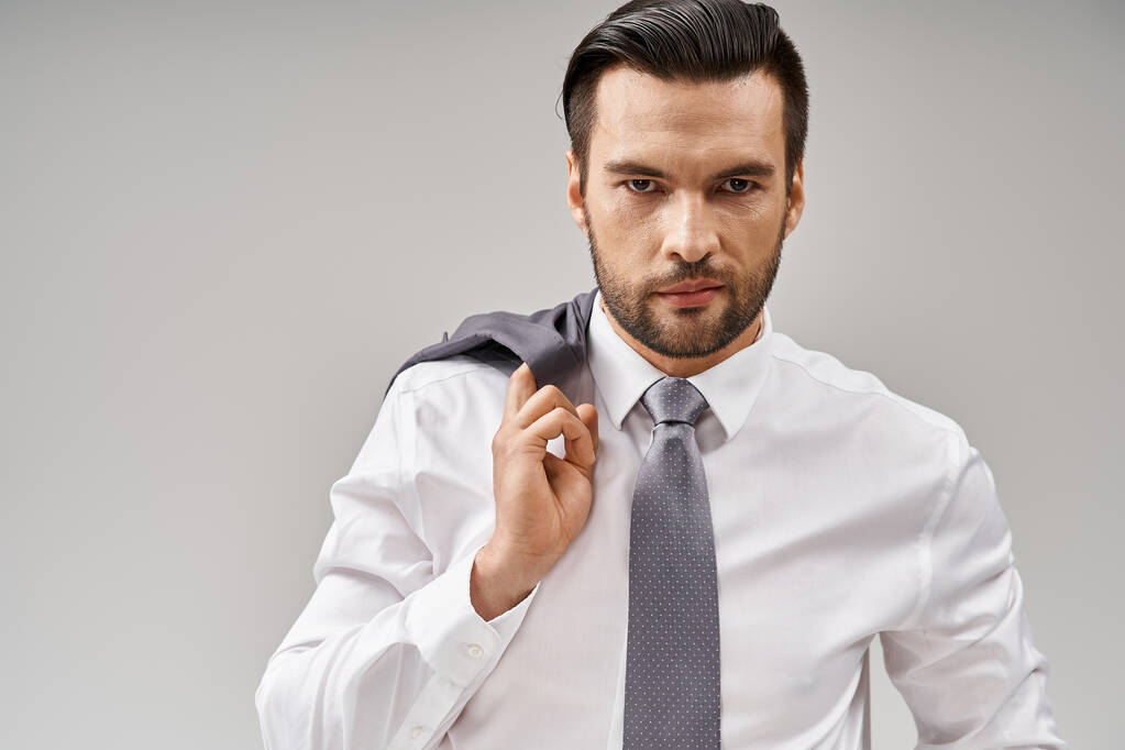 retrato del hombre de negocios de unos 30 años con una chaqueta de cerdas sobre el hombro mientras está de pie sobre gris - Foto, imagen