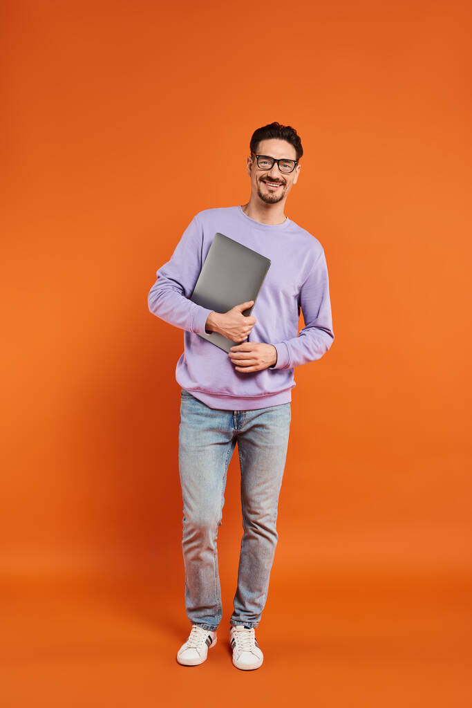 повна довжина бородатого чоловіка в окулярах і фіолетовий светр тримає ноутбук на помаранчевому фоні - Фото, зображення