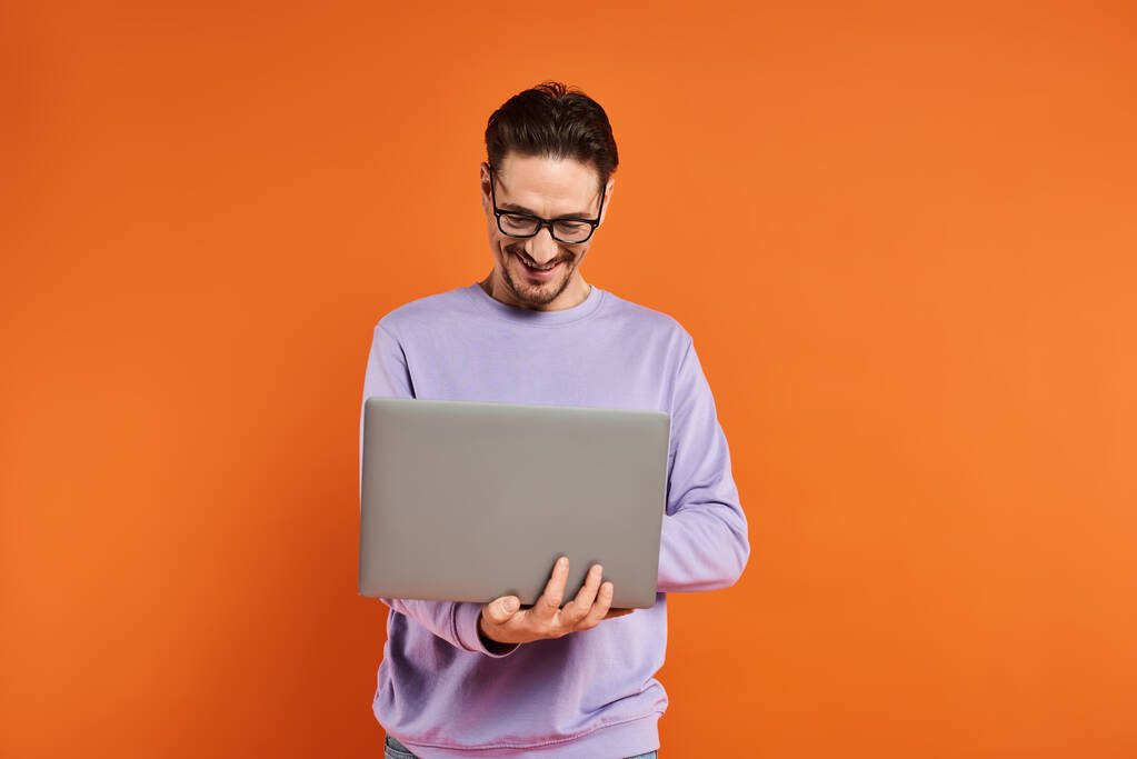 bärtiger Mann mit Brille und lila Pullover mit Laptop auf orangefarbenem Hintergrund, Fernarbeit - Foto, Bild