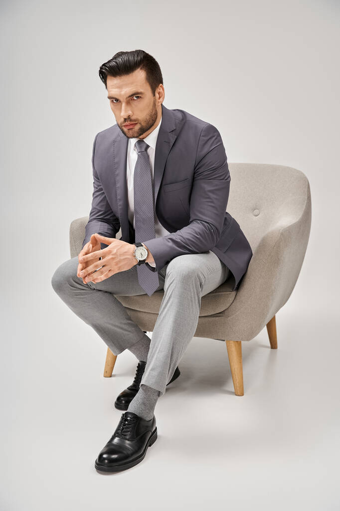 Selbstbewusster und gutaussehender Geschäftsmann im eleganten Anzug sitzt auf einem bequemen Sessel vor grauem Hintergrund - Foto, Bild