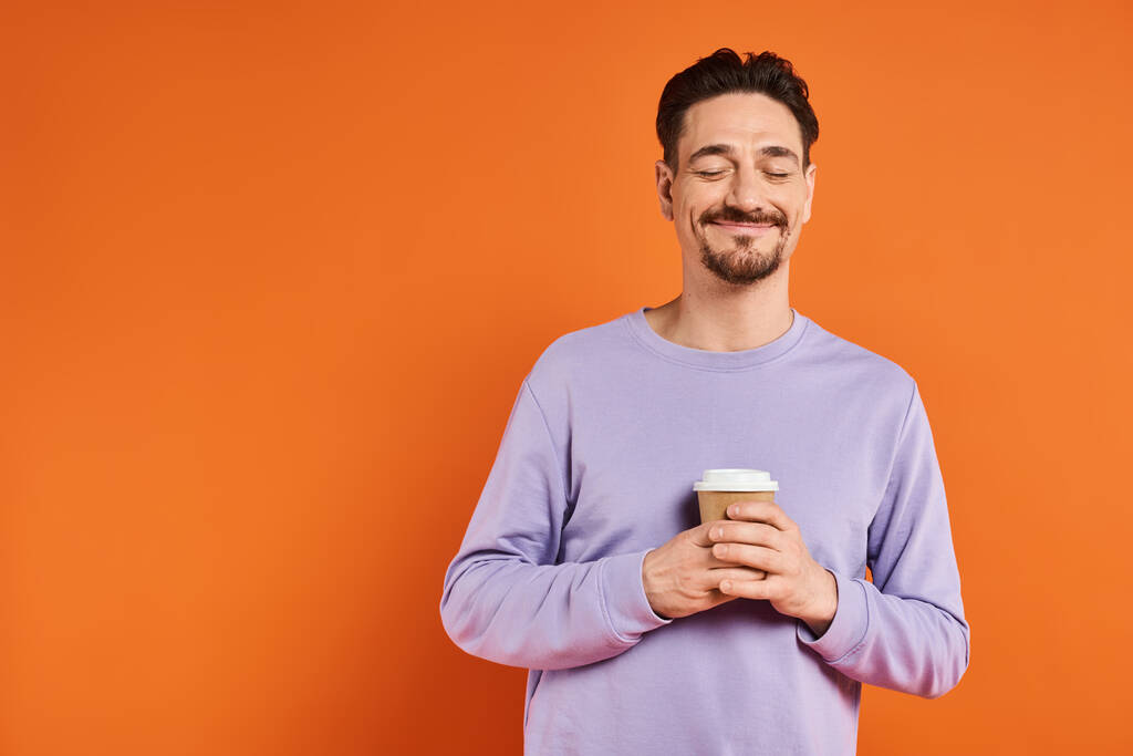 задоволений і бородатий чоловік тримає паперову чашку з кавою, щоб піти на помаранчевий фон, насолода - Фото, зображення