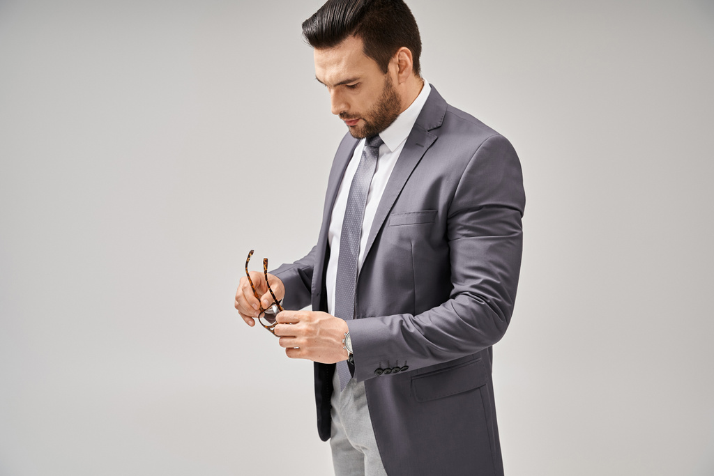 красивий бізнесмен в формальному одязі тримає окуляри і стоїть на сірому фоні, елегантність - Фото, зображення