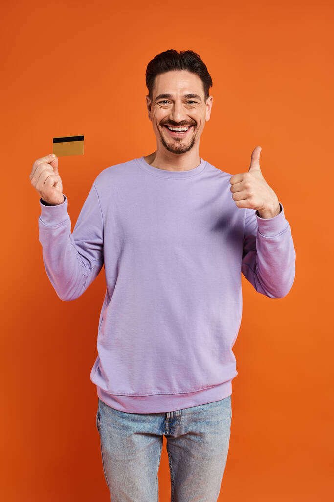 fröhlicher Mann mit Bart, Daumen hoch und Kreditkarte auf orangefarbenem Hintergrund, wie - Foto, Bild