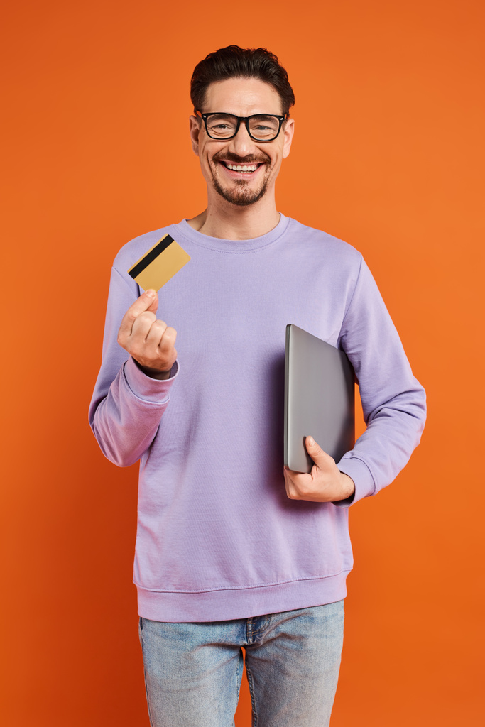 hombre alegre en gafas de pie con tarjeta de crédito y portátil sobre fondo naranja, compras en línea - Foto, imagen