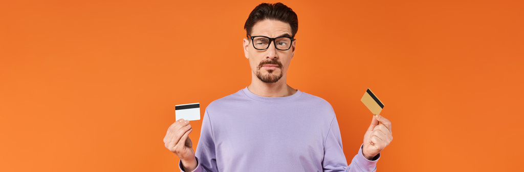skeptický muž v brýlích a fialovém svetru porovnávající dvě kreditní karty na oranžovém pozadí, banner - Fotografie, Obrázek