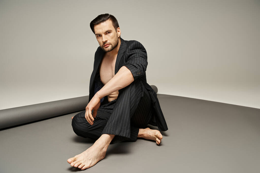 модний і красивий чоловік з голими грудьми і ногами позує в костюмі з шпильками на сірому фоні - Фото, зображення