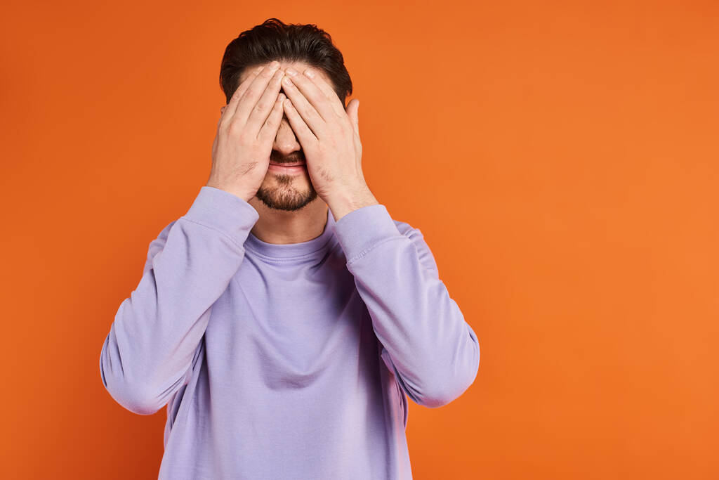 man with beard in purple casual sweater hiding face with hands on orange background, playful mood - Photo, Image