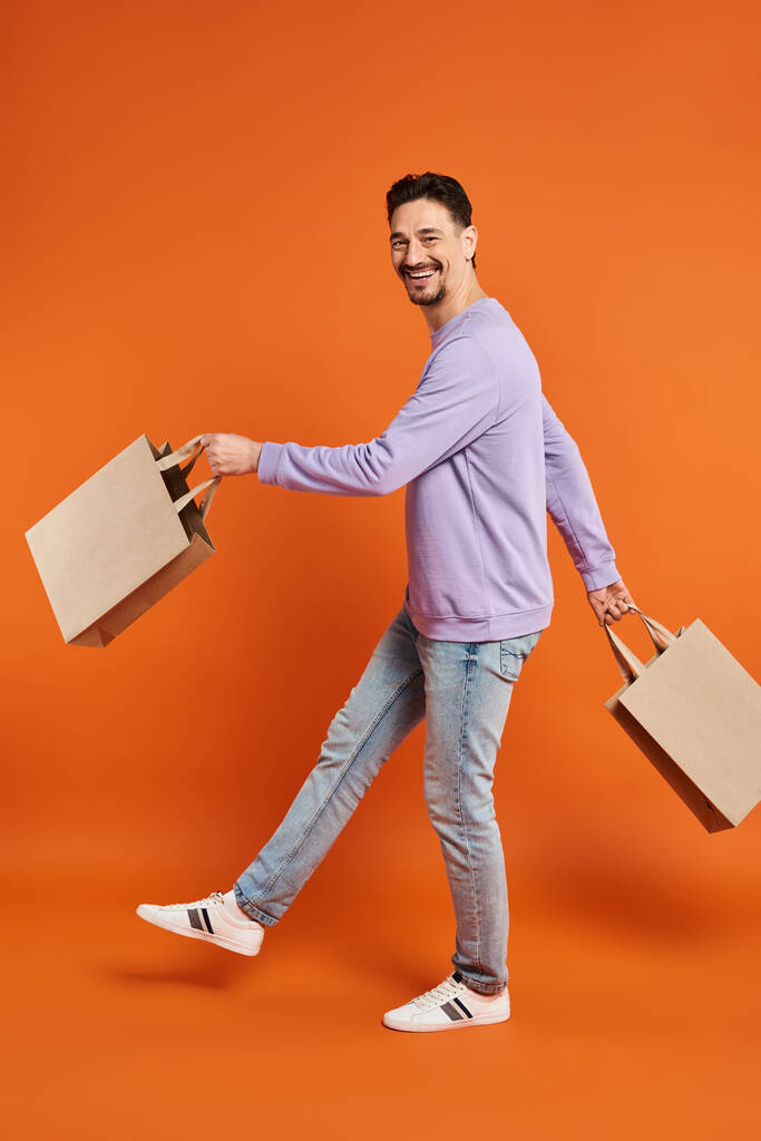 オレンジ色の背景のショッピングバッグで歩くカジュアルな服装で幸せなひげを被った男の全長 - 写真・画像
