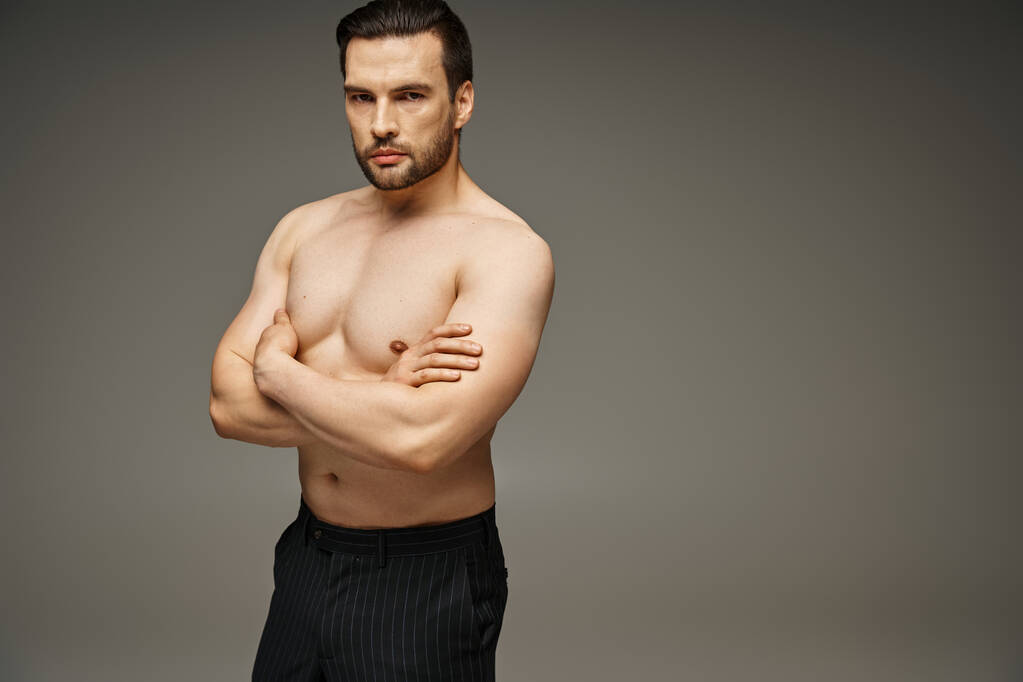 Porträt eines brünetten und gut aussehenden Mannes mit nackter Brust, der mit verschränkten Armen auf grauem Hintergrund posiert - Foto, Bild