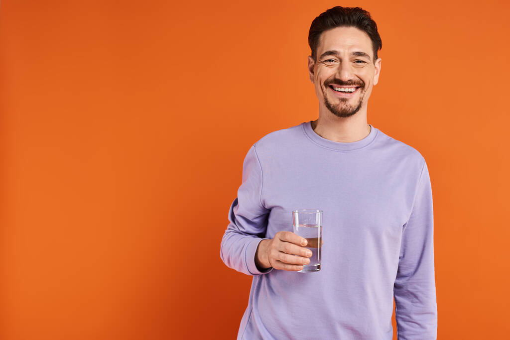 felice uomo barbuto in felpa viola con un bicchiere d'acqua su sfondo arancione, idratazione - Foto, immagini