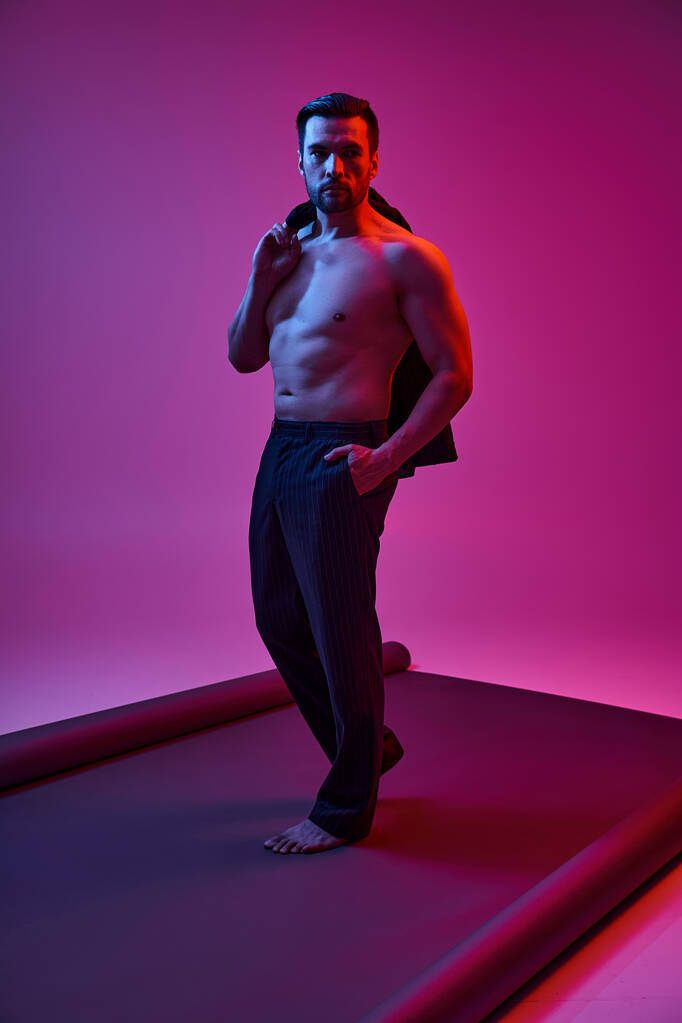 красивий чоловік з голими грудьми і щетиною тримає піджак, позує з рукою в кишені на фіолетовому - Фото, зображення