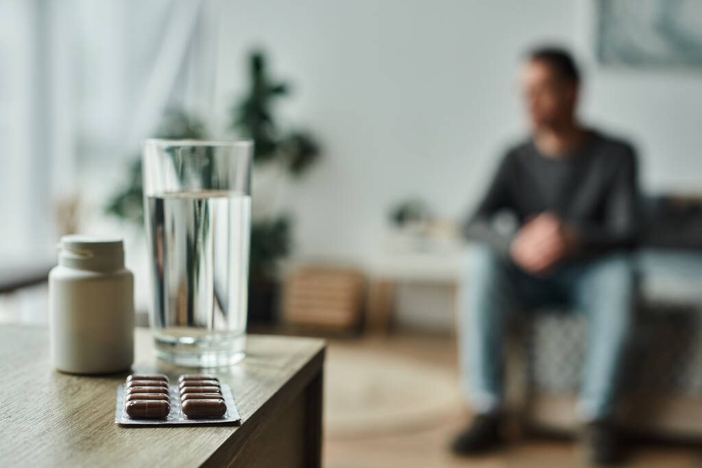 medicação em garrafa e blister pack perto de vidro de água na mesa e homem borrado no fundo - Foto, Imagem