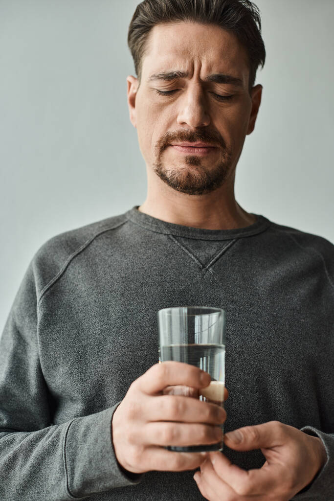 бородатый человек гримасничает, страдая от головной боли и держа стакан воды дома, нездоровым - Фото, изображение