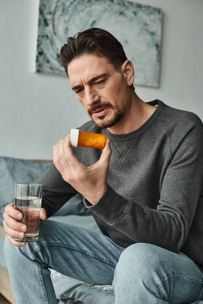 szakállas férfi szürke pulcsiban ráncolja a homlokát, és kezében pohár vizet, miközben nézi palack tabletták - Fotó, kép