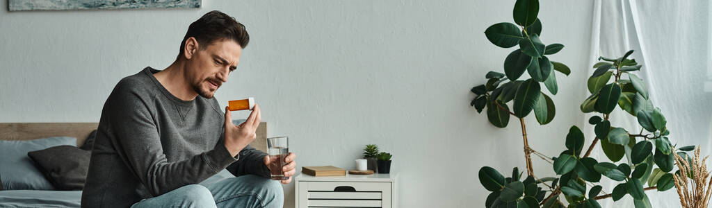 pancarta del hombre barbudo sosteniendo un vaso de agua mientras mira la medicación y se sienta en la cama - Foto, imagen