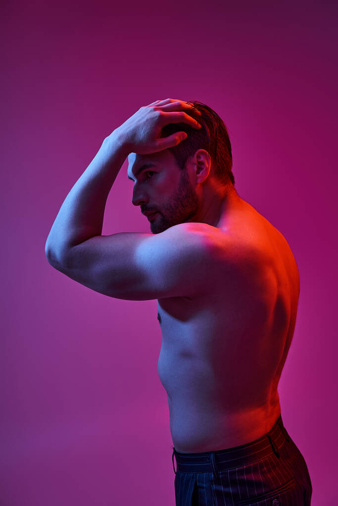 seductive man with bare chest posing with hand near hair on purple backdrop with red lights - Photo, Image