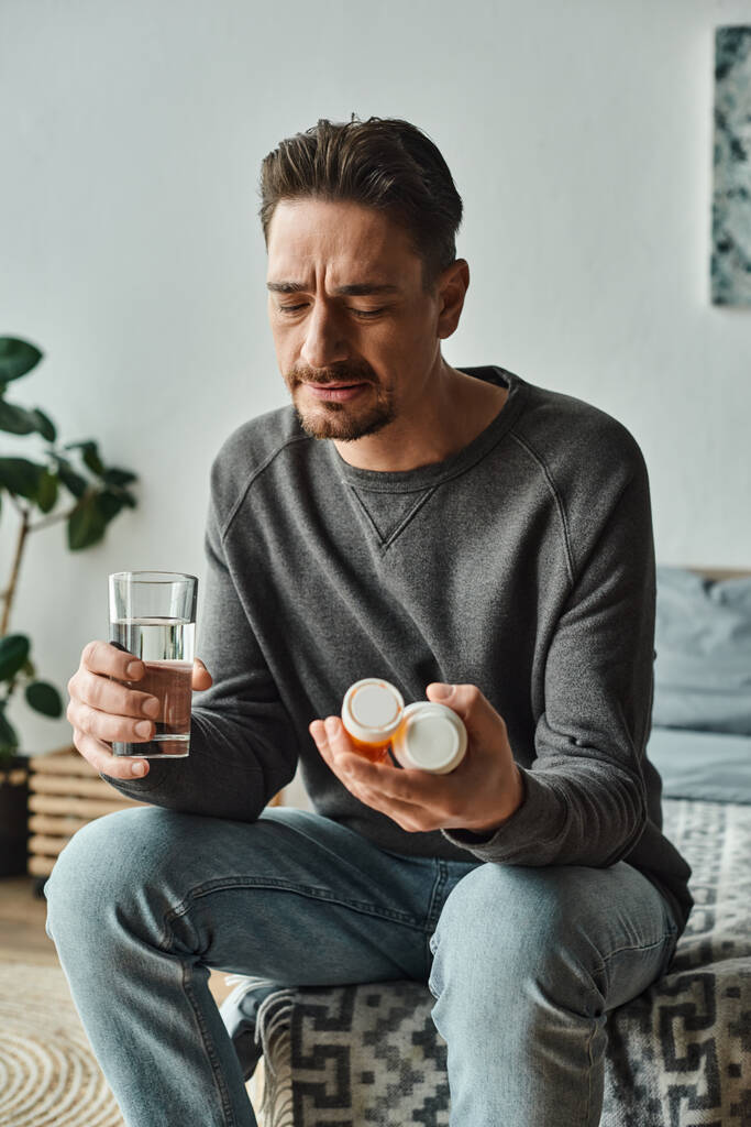 vousatý muž drží sklenici vody, zatímco se dívá na léky v lahvích a sedí na posteli - Fotografie, Obrázek