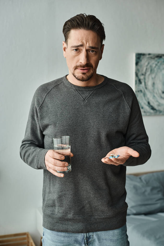 бородатий чоловік дивиться на камеру, тримаючи в руках склянку води і ліки, лікування - Фото, зображення