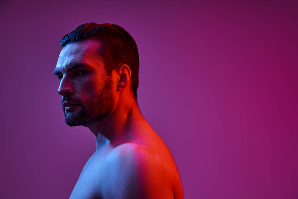 портрет сексуального и мускулистого человека с щетиной, позирующей на фиолетовом фоне с красными и синими огнями - Фото, изображение
