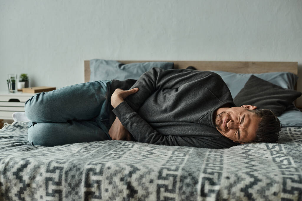 bärtiger Mann, der auf dem Bett liegt und vor Schmerzen die Stirn runzelt, während er unter Bauchschmerzen, Grimassen und Schmerzen leidet - Foto, Bild