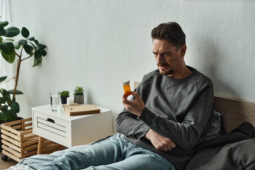 σκεπτικός γενειοφόρος άνδρας εξέταση μπουκάλι με χάπια, ενώ κάθεται στο κρεβάτι στην κρεβατοκάμαρα, σχέδιο θεραπείας - Φωτογραφία, εικόνα