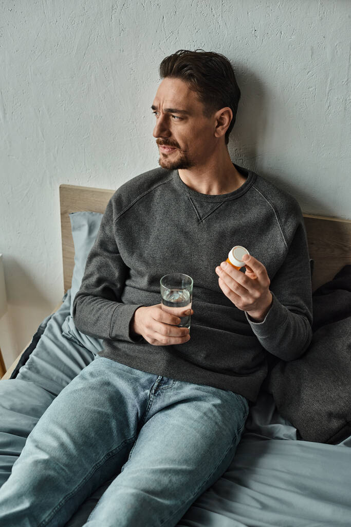 homem pensativo em suéter segurando vidro de água e garrafa com pílulas no quarto, tratamento - Foto, Imagem