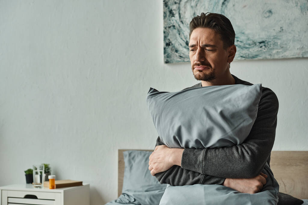 homem barbudo sentindo desconforto e segurando travesseiro enquanto sentado na cama em casa, paciente triste - Foto, Imagem