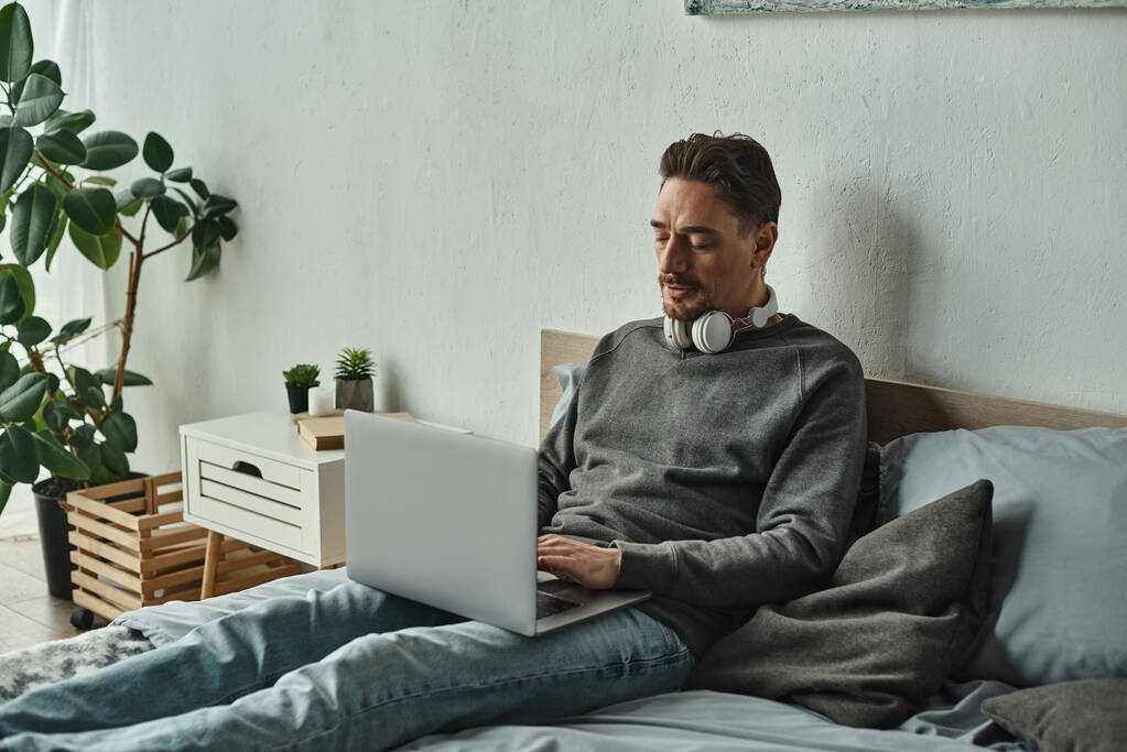 бородатый человек в беспроводных наушниках с помощью ноутбука, сидя на кровати, внештатная концепция - Фото, изображение