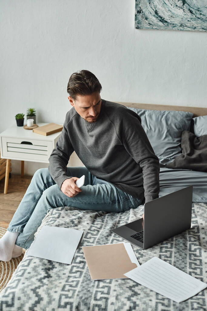 konzentrierter bärtiger Mann mit Smartphone und Laptop in der Nähe von Dokumenten auf dem Bett, Fernarbeit - Foto, Bild