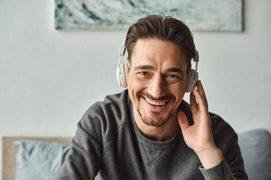 vidám férfi vezeték nélküli fülhallgatóval és szürke alkalmi pulóverben zenét hallgat és kamerába néz - Fotó, kép