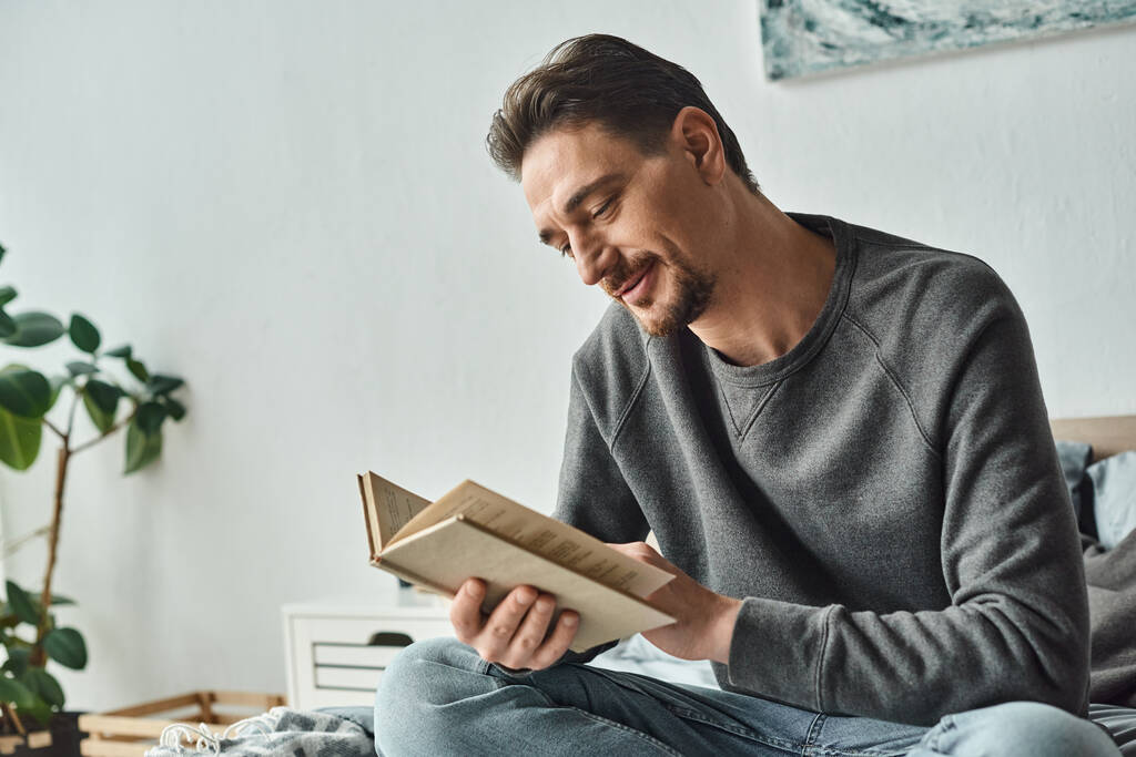 elégedett férfi szürke alkalmi jumper olvasókönyv, miközben pihentető hétvégén a hálószobában, szabadidő - Fotó, kép