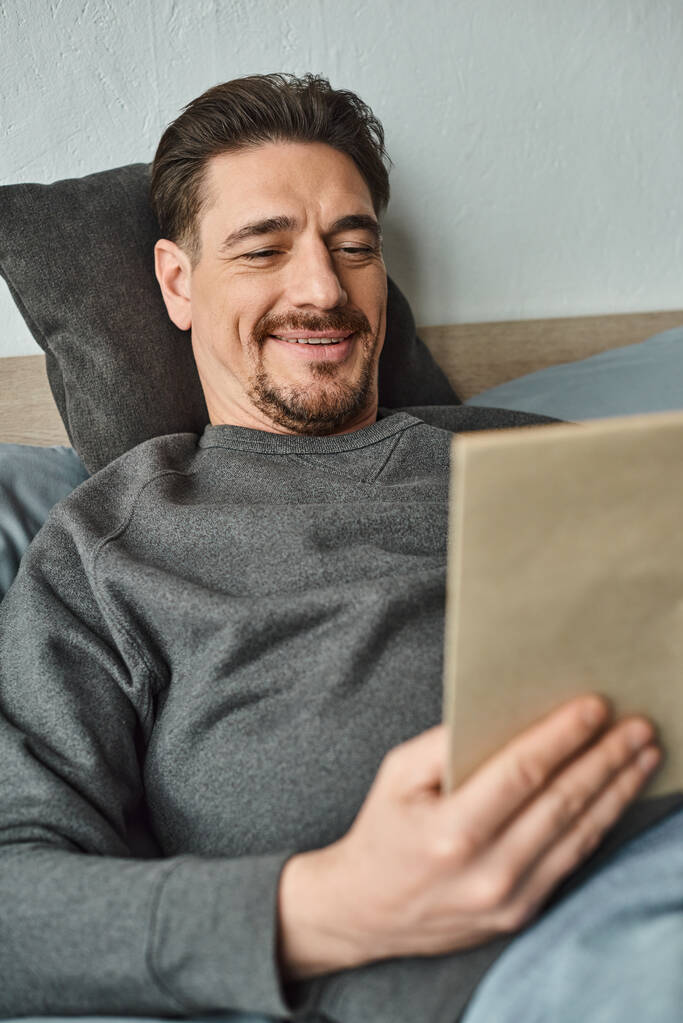 alegre hombre en jumper casual libro de lectura mientras se relaja en fin de semana en el dormitorio, concepto de ocio - Foto, imagen