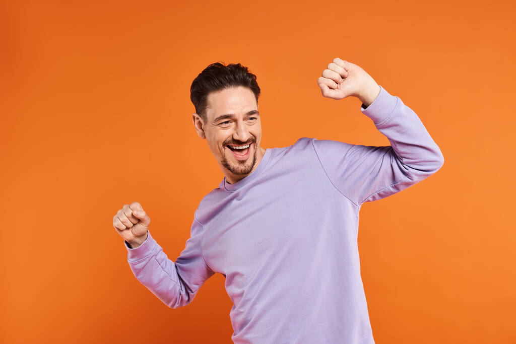 retrato de hombre excitado en suéter morado bailando sobre fondo naranja, persona encantada y divertida - Foto, Imagen