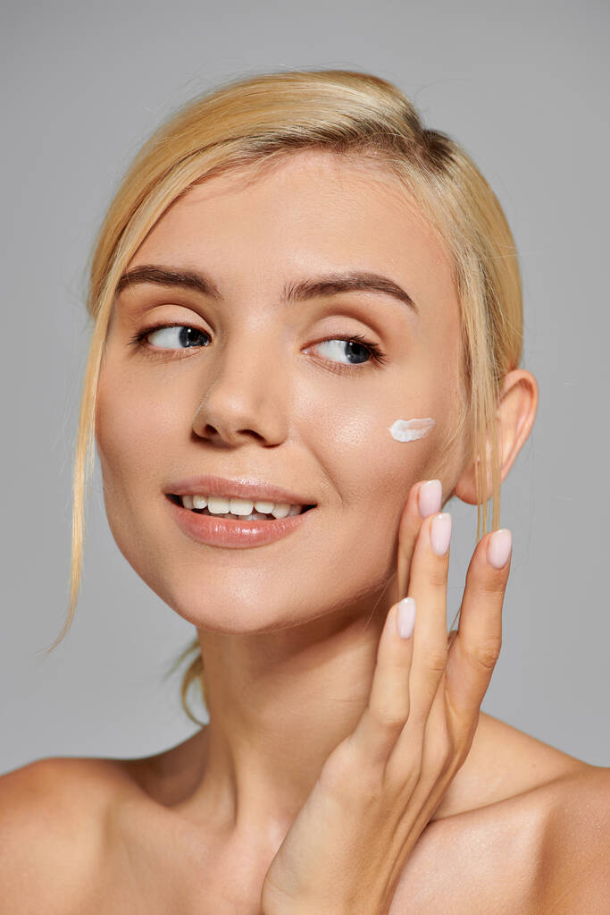 portrait pretty girl in her 20s with healthy skin applying cream on face in grey background - Photo, Image