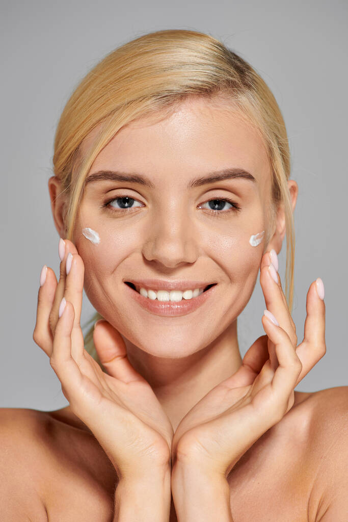 portrait jeune femme souriante avec les mains autour du visage et crème sur les joues sur fond gris - Photo, image