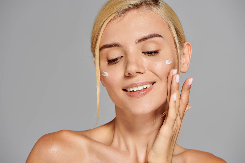 verleidelijke vrouw in de 20 met een gezonde huid blij om crème aan te brengen op haar gezicht in grijze achtergrond - Foto, afbeelding