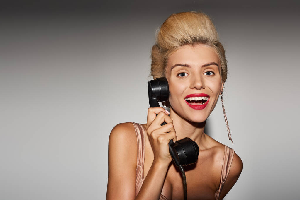 Stilvolles Mädchen in den Zwanzigern mit blonden Haaren greift zum Retro-Handy vor grauem Hintergrund - Foto, Bild
