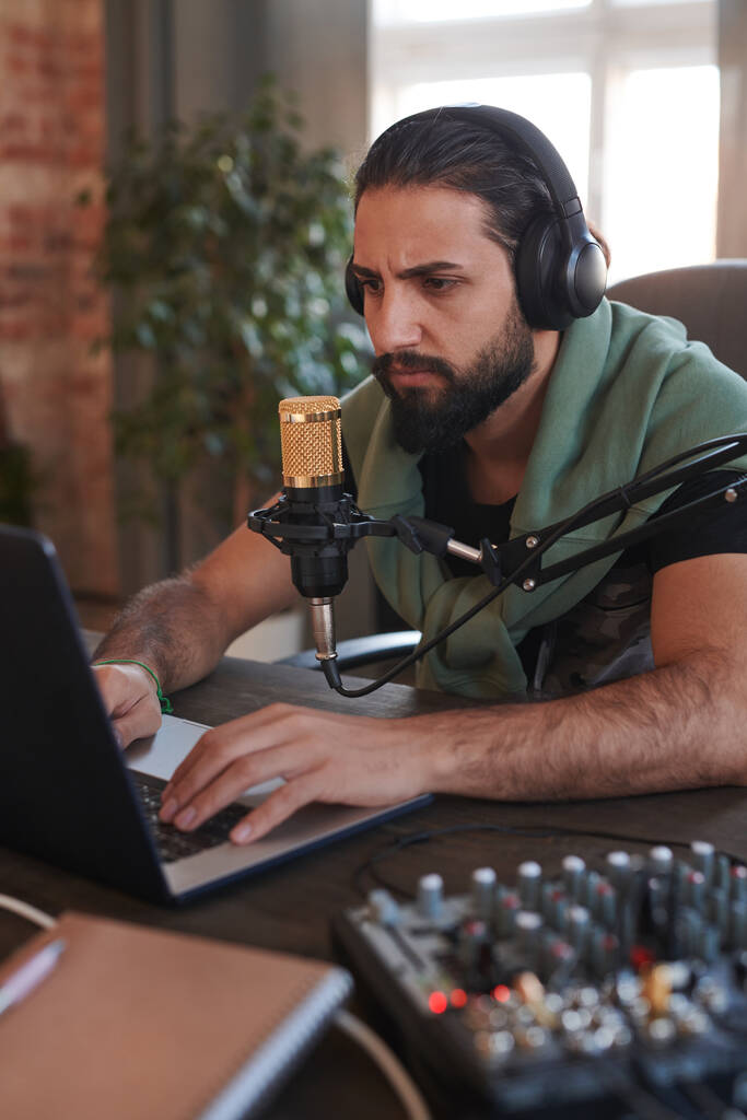 Retrato vertical medio de un hombre guapo y serio con auriculares sentados en el escritorio escribiendo algo en el portátil - Foto, Imagen