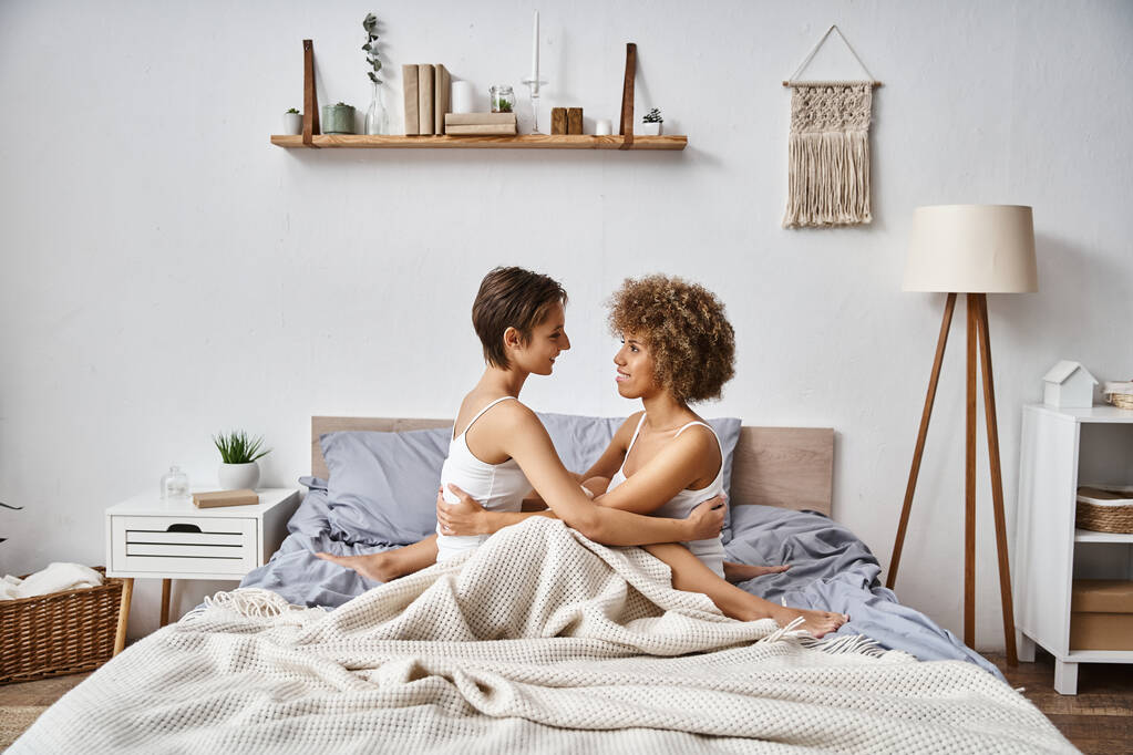 vrolijk multicultureel en jong lesbisch paar knuffelen en kijken naar elkaar in slaapkamer - Foto, afbeelding