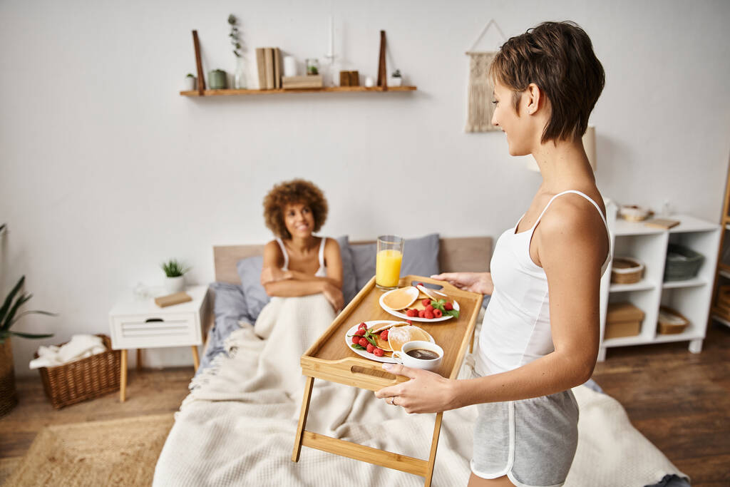счастливая женщина держит поднос с блинами и стаканом апельсинового сока рядом с подругой, завтрак в постель - Фото, изображение