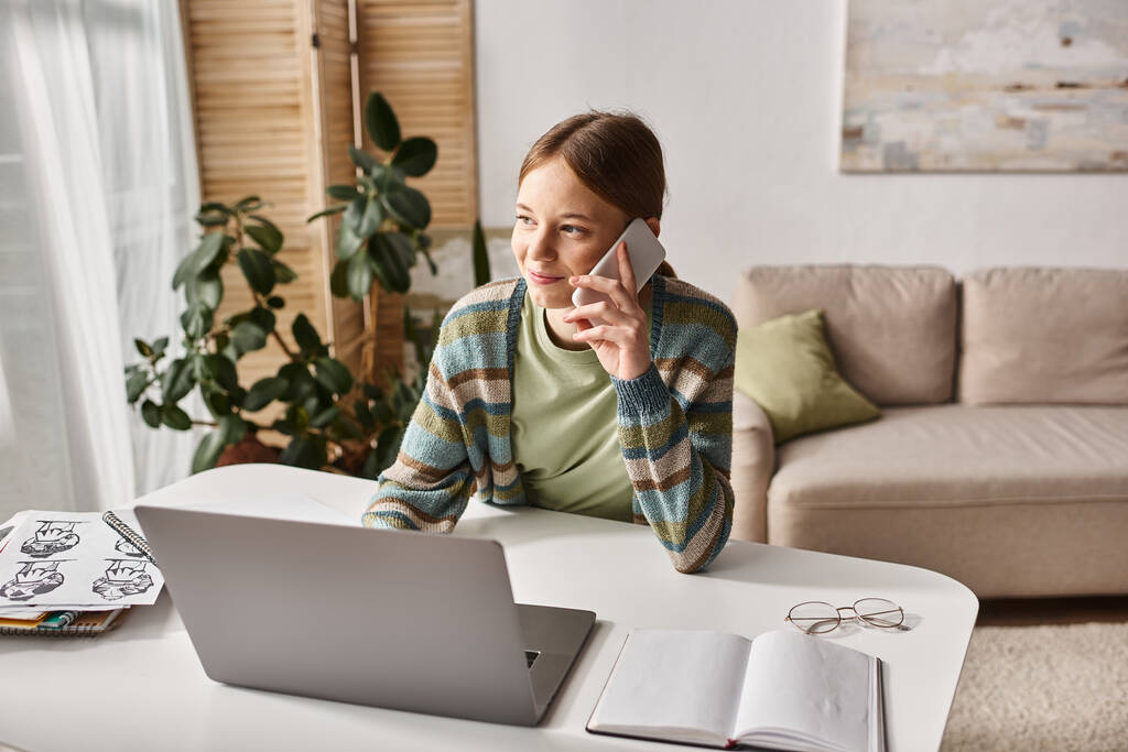 Sonriente adolescente haciendo una llamada telefónica mientras está sentado cerca de la computadora portátil en el escritorio, sesión de estudio electrónico - Foto, imagen