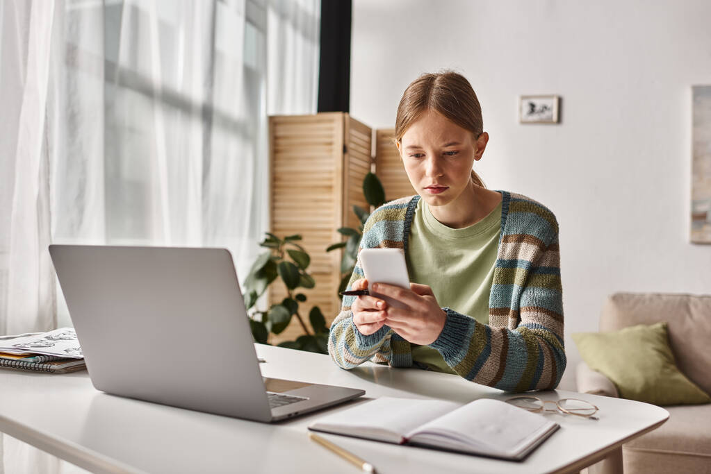 сконцентрированная девочка-подросток с помощью смартфона, сидя рядом с ноутбуком на столе, электронное обучение - Фото, изображение